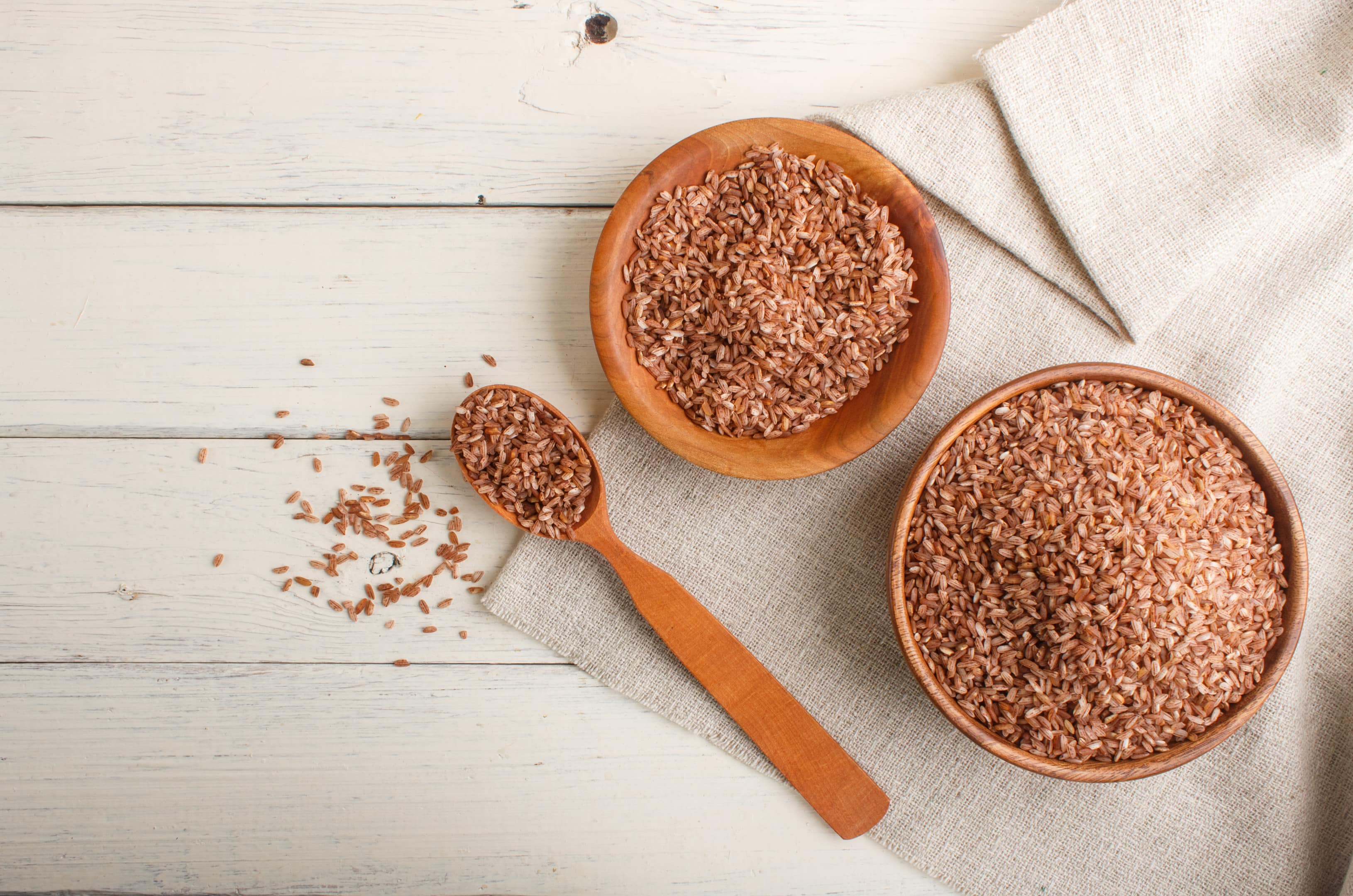 Two wooden bowls with unpolished brown rice with wooden spoon on white wooden background