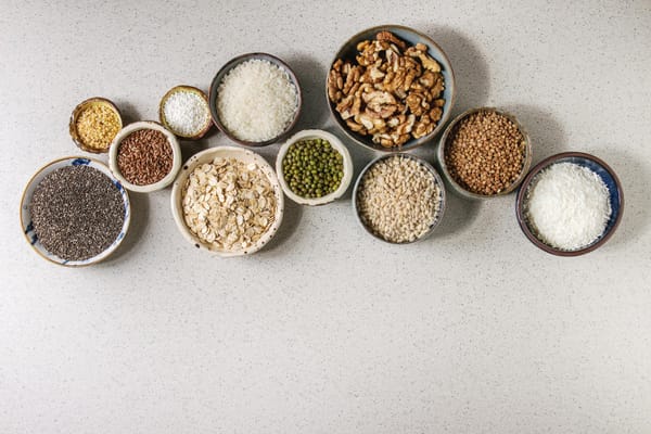 Variety of raw uncooked grains superfood cereal chia, linen, sesame, mung bean, walnuts, tapioca, wheat, buckwheat, oatmeal, coconut, rice in ceramic bowls grey spotted background