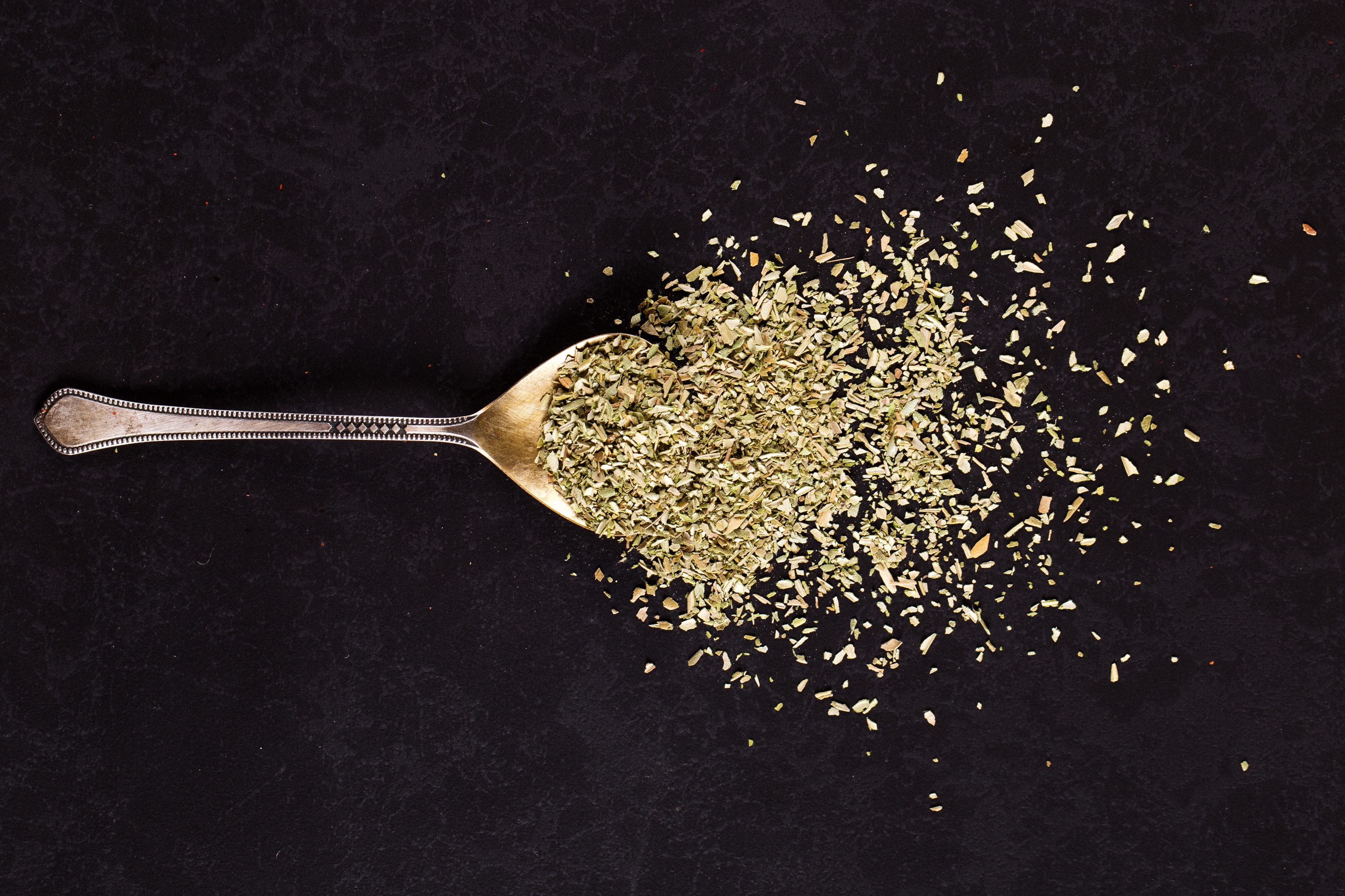 Crushed oregano scattered from iron spoon on black table
