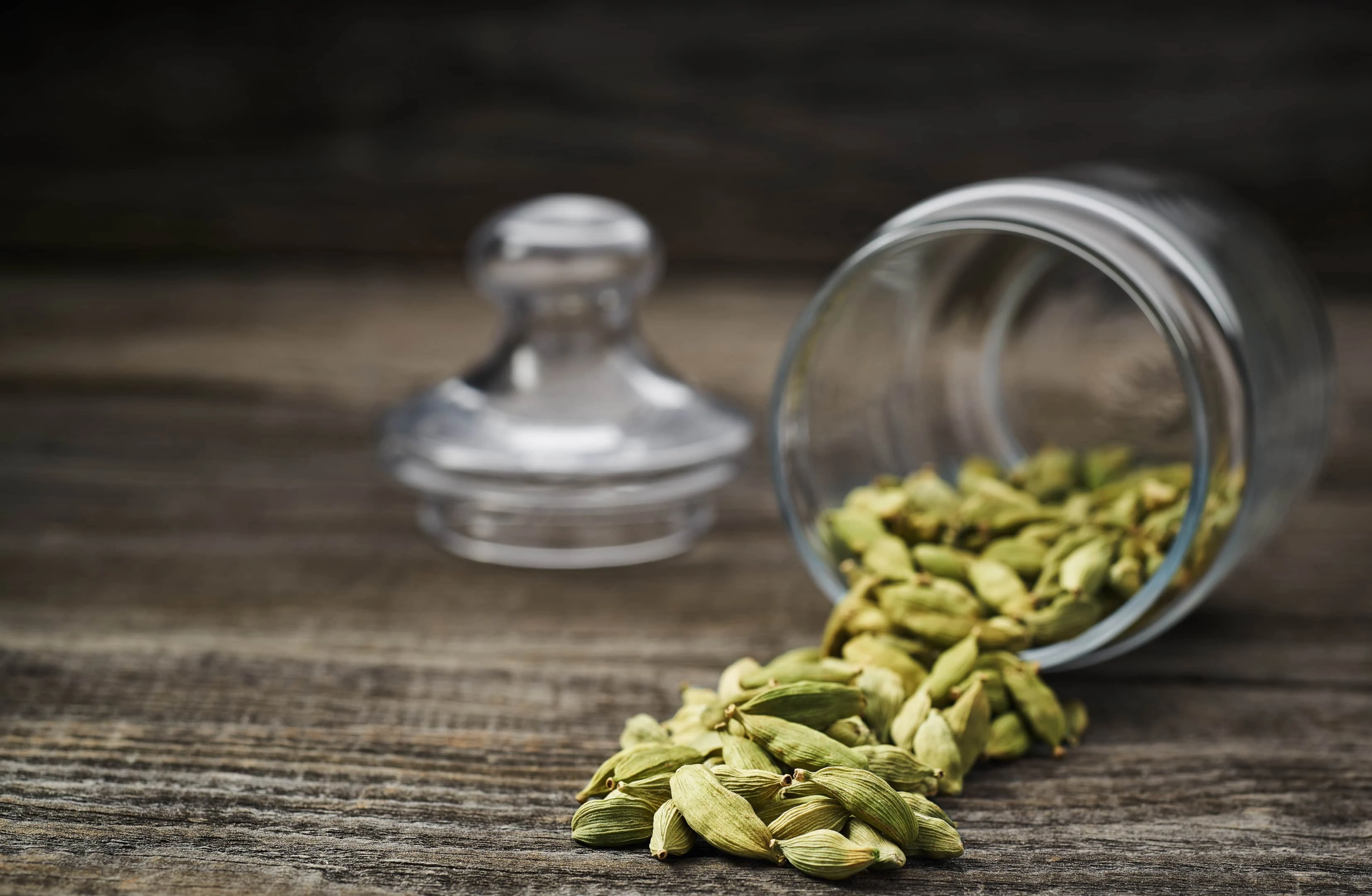 Green cardamom pods spill from glass jar on wooden table