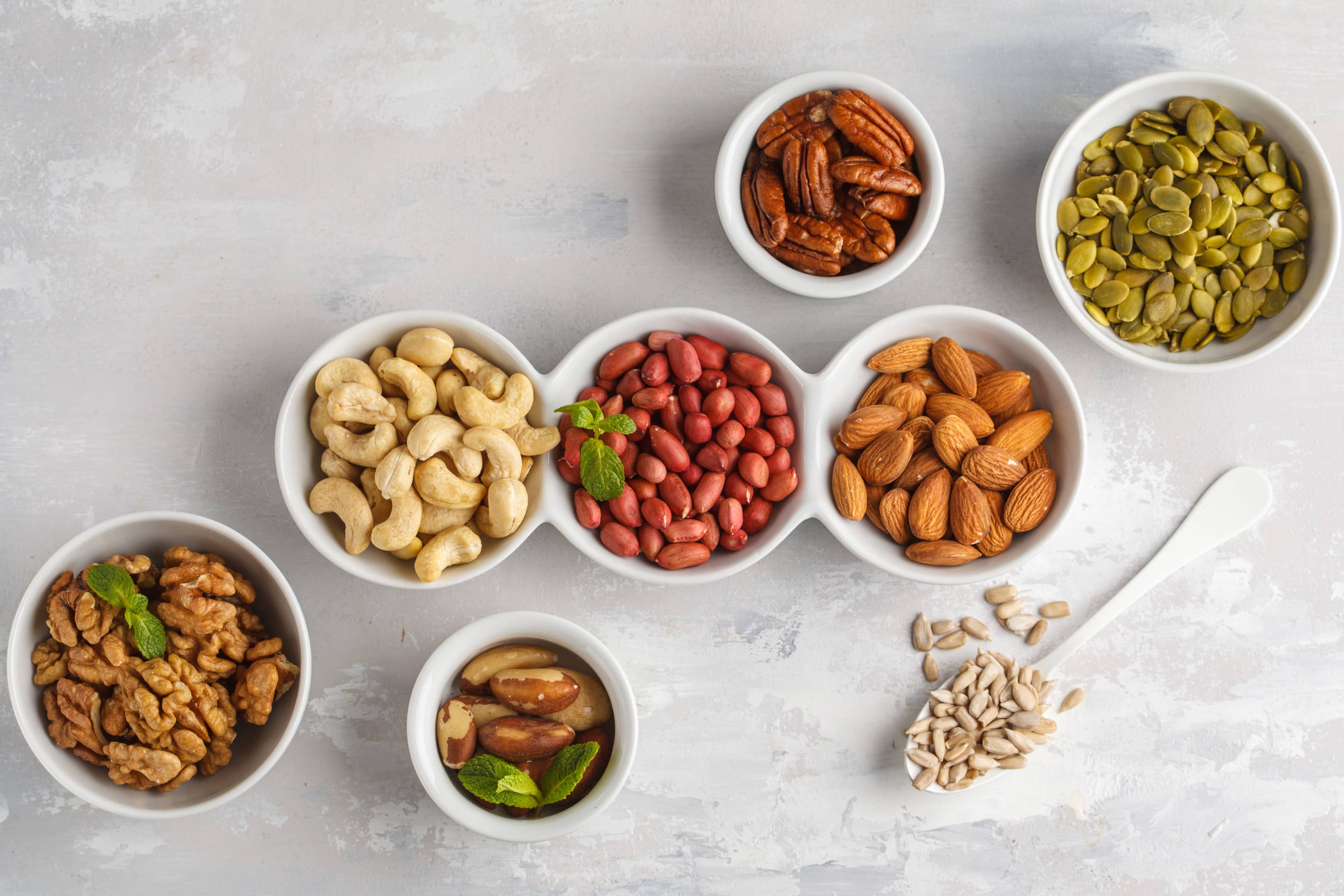 Variety of nuts and seeds in white bowls on gray background