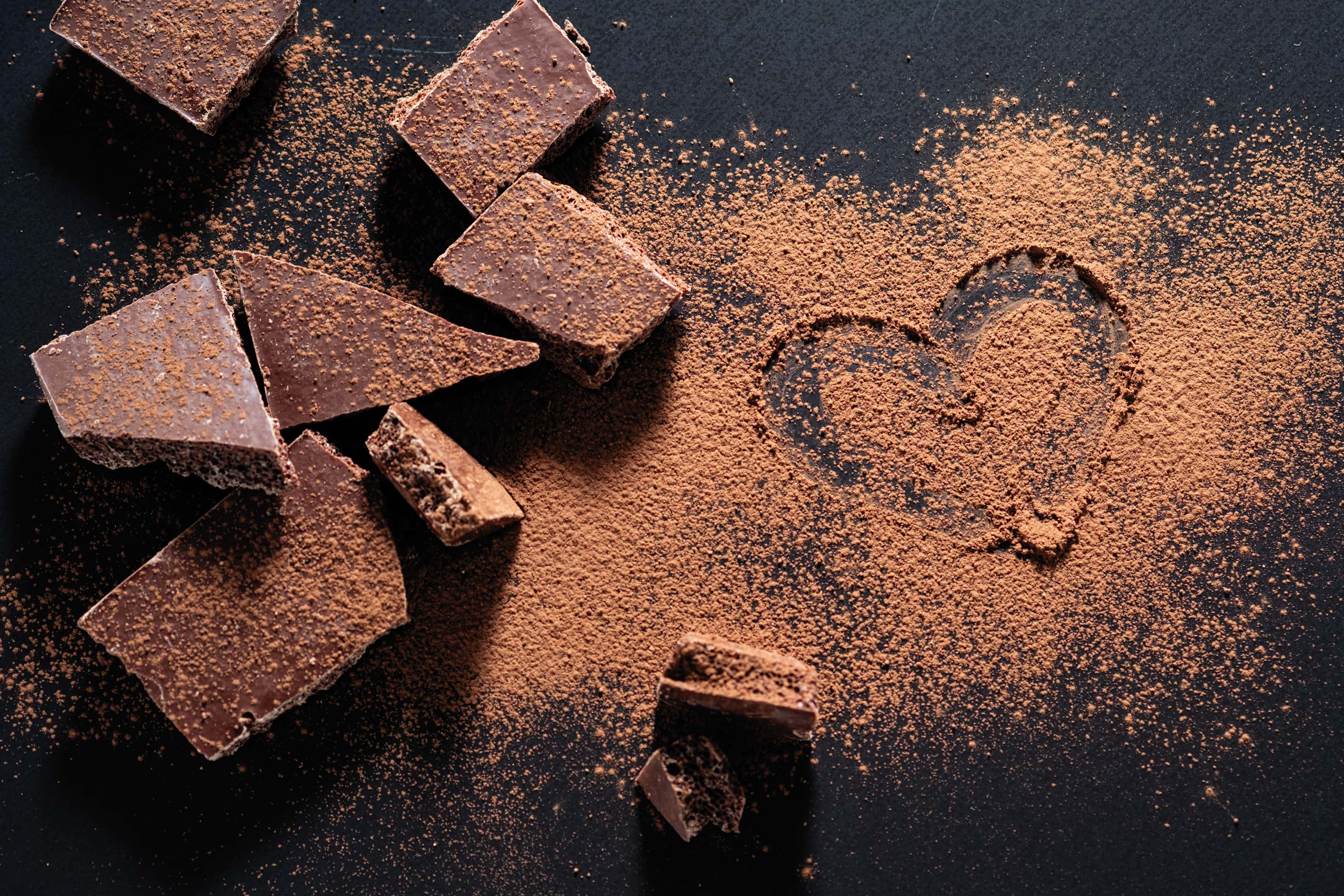 Broken dark chocolate bar with cocoa powder painted heart on black background
