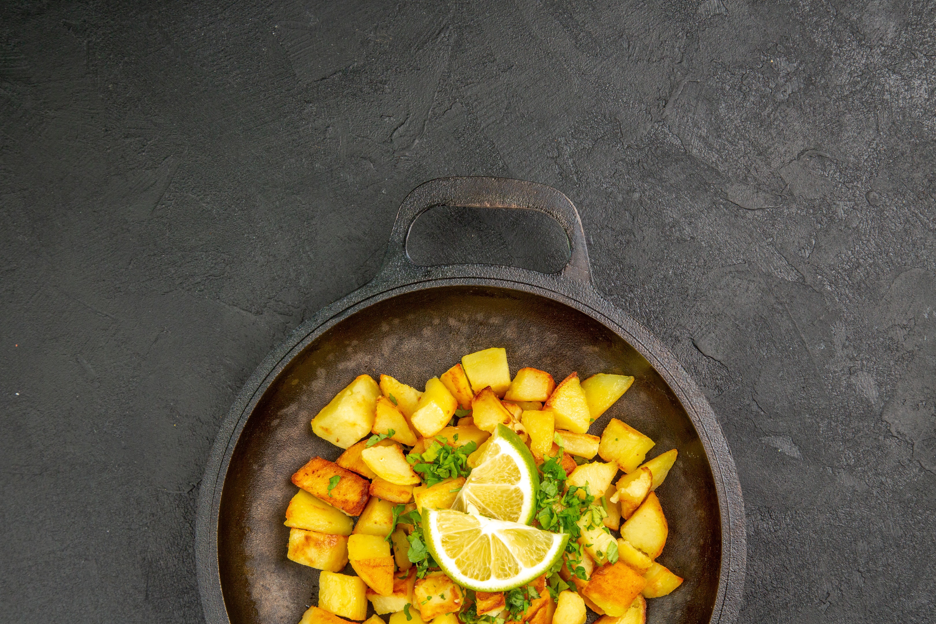 Fried potatoes in a pan with lemon slices on dark surface