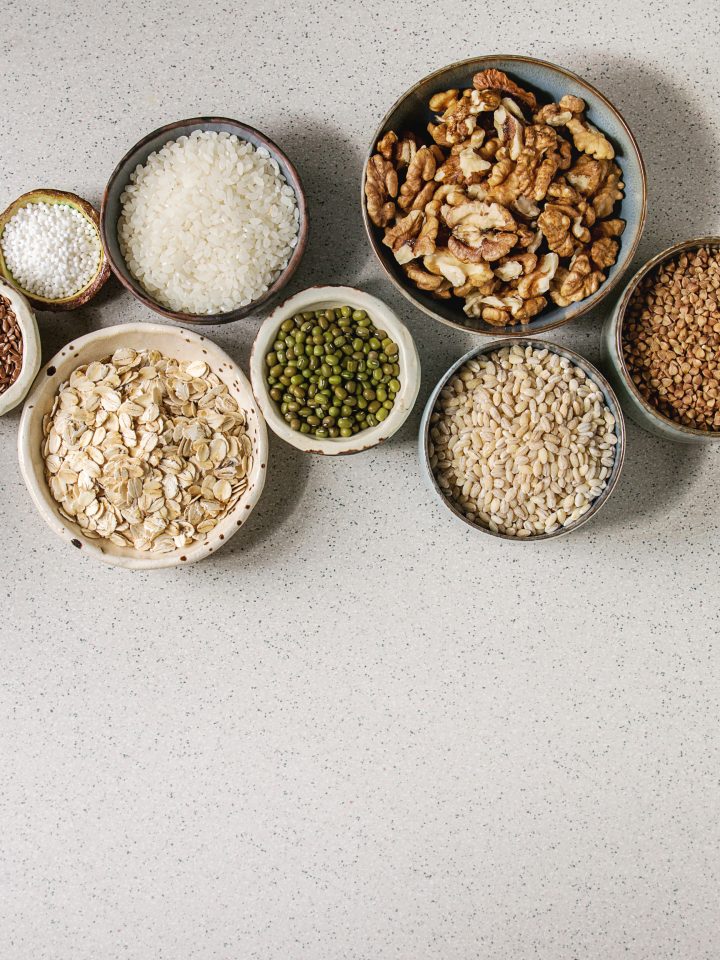 Variety of raw uncooked grains superfood cereal chia, linen, sesame, mung bean, walnuts, tapioca, wheat, buckwheat, oatmeal, coconut, rice in ceramic bowls grey spotted background