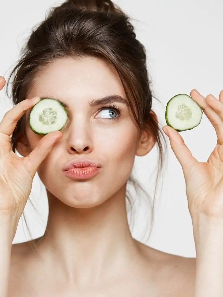 Young beautiful naked girl smiling hiding eye behind cucumber slice over white background